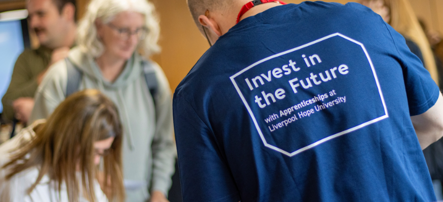 A staff member stood with his back to the camera, wearing a branded T-shirt with Invest in the future with apprenticeships at Liverpool Hope prominent in the image.